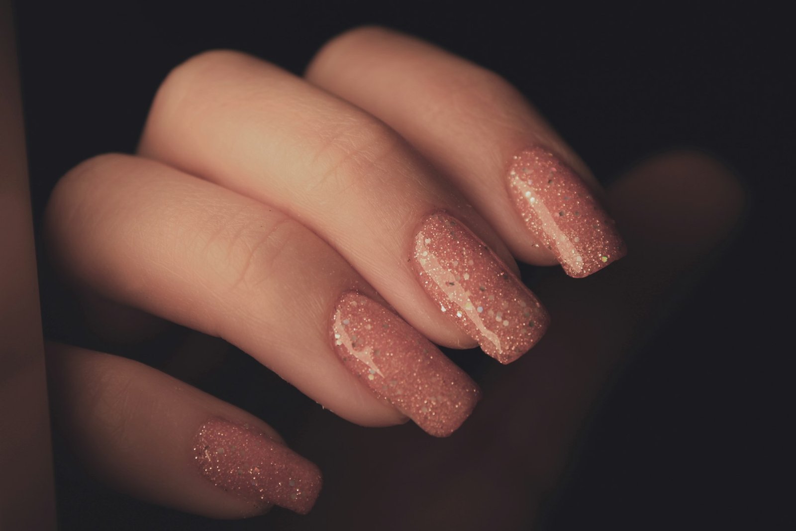 Hands with glittery nude almond nails against a dark background